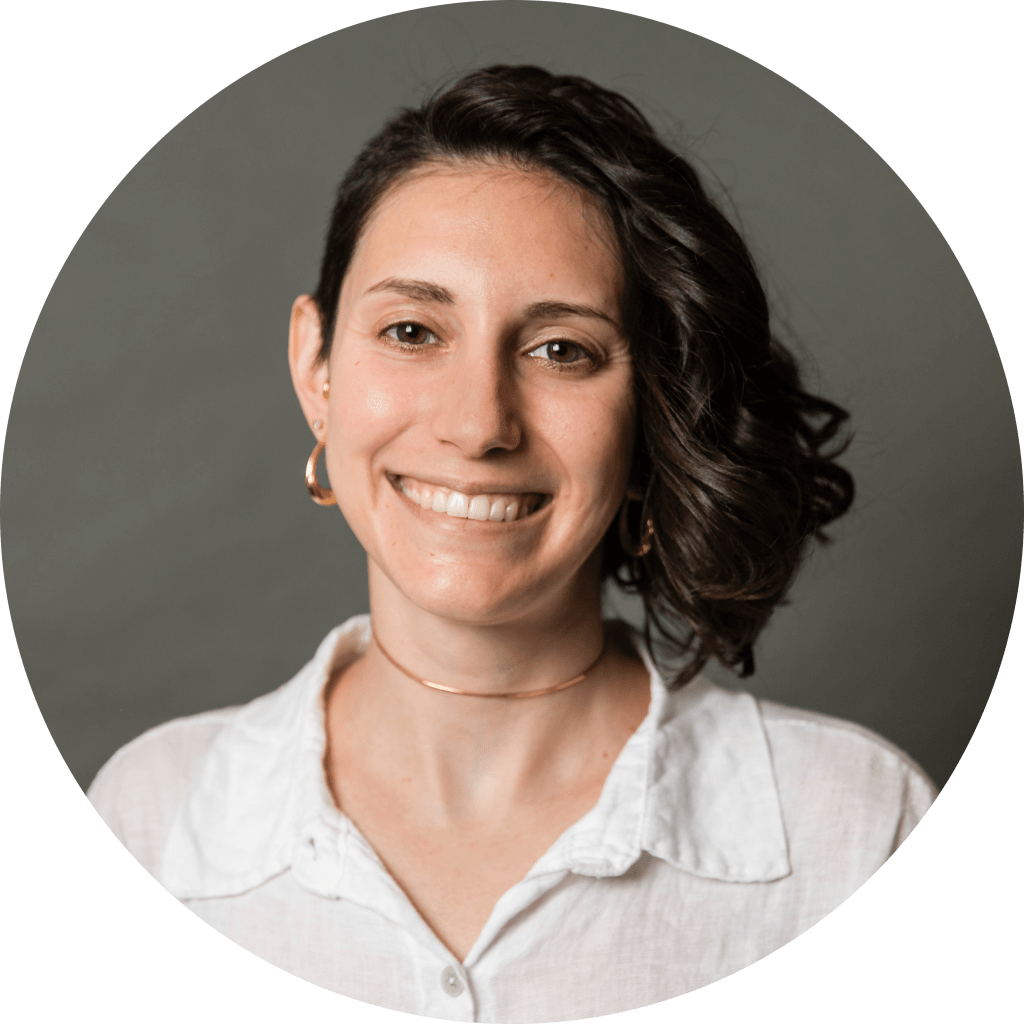 Megan smiles at the camera. She is white with dark brown/black hair that is shaved on one side and curly just past the chin on the other. They are wearing a white collared button down linen shirt, a gold choker, and gold hoop earrings.